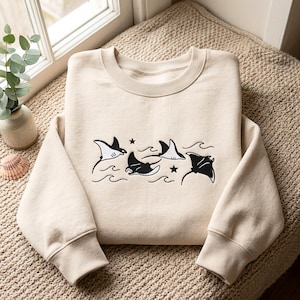 May include: A beige crewneck sweatshirt featuring an embroidered graphic of four black and white manta rays swimming among stylised waves and two small stars. The long-sleeved garment has a ribbed collar, cuffs, and hem, with a clean, simple design.