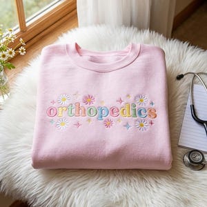 Könnte beinhalten: Ein hellrosa Sweatshirt mit Rundhalsausschnitt und dem Wort orthopedics, das in mehrfarbigen Pastellbuchstaben auf die Brust gestickt ist. Der Schriftzug ist von gestickten weißen und rosa Gänseblümchen sowie kleinen bunten vierzackigen Sternen umgeben.
