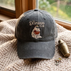 May include: A charcoal grey baseball cap featuring an embroidered graphic of a white cat wearing sunglasses and a small crown, holding a cocktail glass with a cherry. The text above the cat reads THE DESPERADO CLUB in a cream-coloured font.