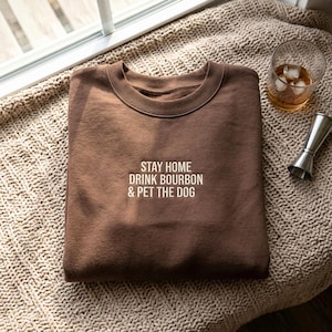 May include: A brown crewneck sweatshirt featuring the embroidered white text STAY HOME DRINK BOURBON & PET THE DOG centered on the chest. The garment has a ribbed neckline and long sleeves, presented in a folded position with a clear glass of amber liquid.