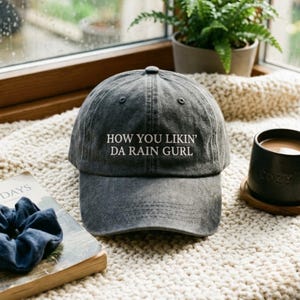 May include: A charcoal gray baseball cap with a distressed finish featuring the white embroidered text HOW YOU LIKIN' DA RAIN GURL centered on the front panel. The cap has a structured crown, a curved brim, and stitched eyelets for ventilation.