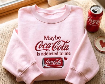 May include: A light pink crewneck sweatshirt featuring red embroidered text that reads Maybe Coca-Cola is addicted to me, accompanied by a small red heart and an embroidered graphic of a red Coca-Cola soda can with white lettering.