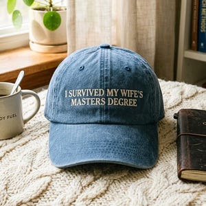 Peut inclure: Une casquette de baseball en jean bleu avec le texte blanc brodé I SURVIVED MY WIFE'S MASTERS DEGREE centré sur le panneau avant. La casquette présente une calotte structurée, une visière incurvée et des coutures visibles sur le tissu.