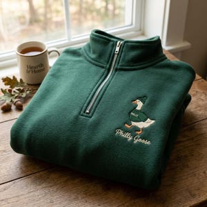 Pode incluir: Um suéter de fleece verde-escuro com zíper de um quarto, apresentando um bordado de um ganso branco vestindo um suéter e um gorro verdes. O ganso tem a inscrição Philly, e o texto Philly Goose está bordado abaixo do gráfico.