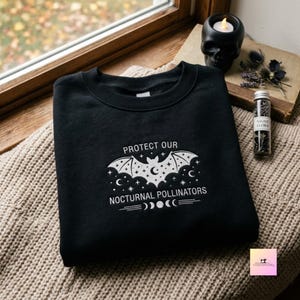 Peut inclure: Un sweat-shirt noir à col rond avec une broderie blanche représentant une chauve-souris au motif étoilé, le texte "PROTECT OUR NOCTURNAL POLLINATORS" et un cycle de phases lunaires centré sur la poitrine du vêtement.
