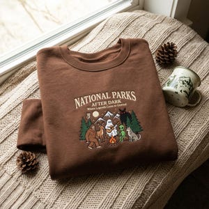 May include: A brown crewneck sweatshirt featuring the text "NATIONAL PARKS AFTER DARK Where Legends Come to Unwind" above a graphic of a campfire, a ghost playing guitar, a Bigfoot, an alien, a deer, a wolf, an owl, and mountains.