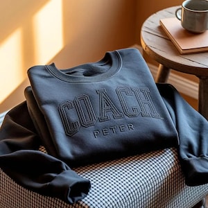 May include: A dark charcoal gray long-sleeve crewneck sweatshirt featuring the word COACH in large, bold, embroidered block lettering across the chest, with the name PETER embroidered in a smaller, simpler font directly underneath the main text.