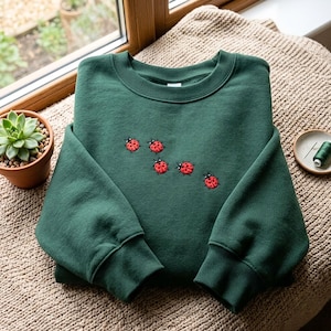 Può includere: Una felpa girocollo color verde bosco scuro con un motivo ricamato di cinque piccole coccinelle rosse a pois neri sparse al centro del petto. Il capo presenta maniche lunghe con polsini e colletto a costine.