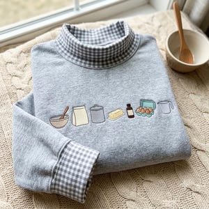 May include: A heather gray sweatshirt featuring a mock neck collar and sleeve cuffs with a gray and white gingham pattern. The front displays an embroidered line of baking ingredients including a bowl, flour bag, canister, butter, extract, eggs, and a pitcher.