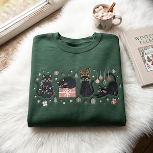 Op de afbeelding: Een donkergroene sweater met ronde hals en een borduursel van vier zwarte katten. De katten zijn afgebeeld met kerstverlichting, een cadeau, rendiergeweien en kerstballen, omringd door kleine sneeuwvlokken en cadeautjes.