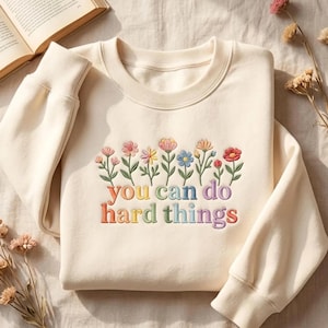 May include: A cream-colored long-sleeve sweatshirt featuring an embroidered design of colorful wildflowers across the chest. Below the flowers, the text "you can do hard things" is embroidered in a multi-colored gradient font with a rounded neckline.