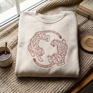 Pode incluir: Uma camisola de gola redonda cor de creme com um bordado vermelho escuro de dois leopardos em formação circular. Os leopardos são representados com manchas detalhadas e bocas abertas, virados um para o outro num padrão fluido e contínuo.