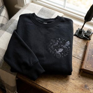 May include: A black crewneck sweatshirt featuring an embroidered design on the left chest. The embroidery depicts three grey dragons surrounded by white stars, constellations, and a crescent moon, all arranged in a circular pattern.