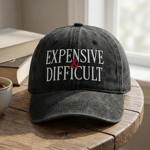 Puede incluir: Una gorra de béisbol gris carbón con un acabado desgastado. La parte frontal presenta un texto bordado en blanco que dice "EXPENSIVE" en la línea superior y "DIFFICULT" en la inferior, separados por un pequeño signo ampersand rojo.