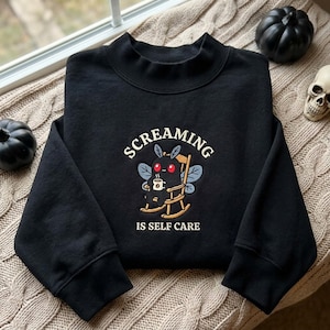 Könnte beinhalten: Ein schwarzes Langarm-Sweatshirt mit einer Stickerei eines schwarzen Nachtfalters mit roten Augen, der in einem hölzernen Schaukelstuhl sitzt und eine weiße Kaffeetasse hält. Der Text lautet oben SCREAMING und unten IS SELF CARE.