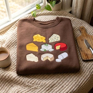 Può includere: Una felpa marrone a girocollo con un ricamo raffigurante vari tipi di formaggio, tra cui svizzero, erborinato, cheddar, feta, una forma con cera rossa, parmigiano, brie e un tronchetto di caprino disposti a griglia.