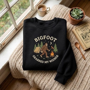 May include: A black crewneck sweatshirt featuring a graphic of Bigfoot roasting a hot dog over a campfire near a tent and pine trees. The text reads BIGFOOT GRABBED MY WIENER in a circular beige font around the central illustration.