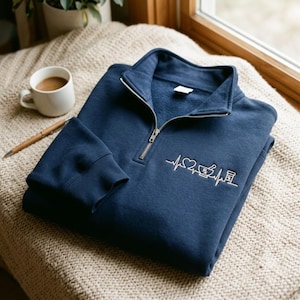 May include: A navy blue quarter-zip sweatshirt featuring an embroidered white graphic on the chest. The design displays a heartbeat line incorporating a heart, a mortar and pestle with an Rx symbol, and a pill bottle icon in a continuous line.