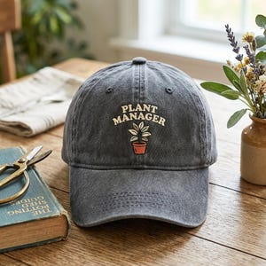 May include: A charcoal grey baseball cap featuring an embroidered design of a potted plant with the text "PLANT MANAGER" arched above it in cream-coloured lettering. The cap has a structured crown, a curved brim, and a classic six-panel construction.