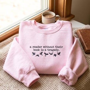 May include: A light pink crewneck sweatshirt featuring black embroidered text that reads, a reader without their book is a tragedy. Below the text are small black embroidered silhouettes of flying dragons arranged in a horizontal row.