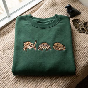 May include: A forest green crewneck sweatshirt featuring an embroidered design of three brown jumping spiders. The left spider is smiling, the middle spider wears black sunglasses, and the right spider is curled up with its eyes closed.