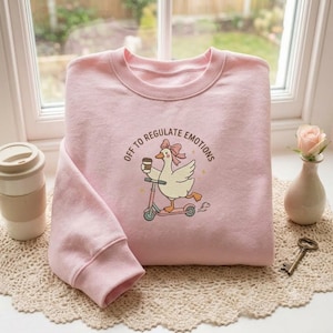 Puede incluir: Una sudadera de cuello redondo rosa claro con un gráfico de un ganso blanco con un lazo rosa, montado en un patinete rosa y sosteniendo una taza de café. El texto sobre el ganso dice "OFF TO REGULATE EMOTIONS" en letras mayúsculas marrones.