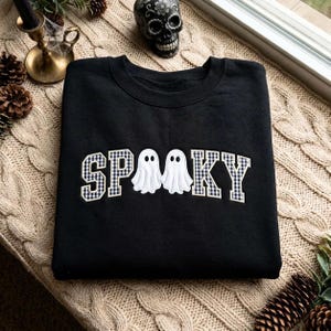 May include: A black crewneck sweatshirt featuring the word SPOOKY embroidered across the chest. The letters S, P, O, K, and Y are filled with a black and white gingham pattern, while two white ghost figures replace the double O in the centre.