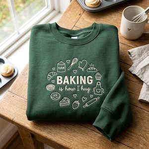 May include: A dark green crewneck sweatshirt featuring a white graphic print of baking-related icons including a whisk, flour container, apron, and various pastries, with the text "BAKING is how I hug" centered among small heart illustrations.