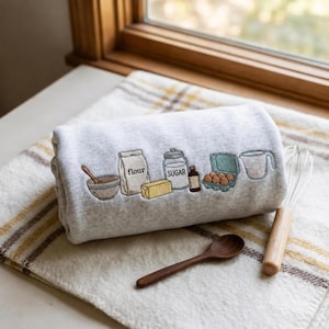 Könnte beinhalten: Ein hellgraues Sweatshirt mit einer Stickerei von Backzutaten, darunter eine Schüssel mit Löffel, eine Tüte mit der Aufschrift flour, ein Stück Butter, ein Glas mit der Aufschrift sugar, eine kleine Flasche, ein Eierkarton und ein Messbecher.