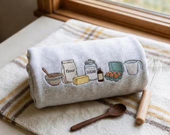 May include: A light gray sweatshirt featuring an embroidered design of baking ingredients including a bowl, a bag labeled flour, a stick of butter, a jar labeled sugar, a bottle, a carton of eggs, and a measuring cup, with a wooden spoon and whisk.