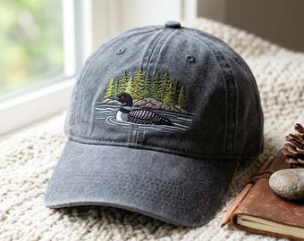 May include: A charcoal gray baseball cap featuring an embroidered design of a black and white loon swimming on water in front of a green pine forest. The cap has a structured crown, a curved brim, and a button at the top center of the panels.