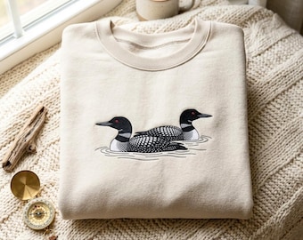May include: A beige crewneck sweatshirt featuring an embroidered graphic of two black and white loons with red eyes swimming on water. The birds are depicted in profile, showcasing detailed plumage patterns and sharp beaks on the light fabric.