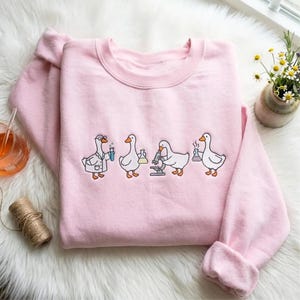 Op de afbeelding: Een lichtroze sweater met ronde hals en een geborduurde afbeelding van vier witte ganzen die wetenschappelijke experimenten uitvoeren. De ganzen dragen laboratoriumjassen, gebruiken een microscoop en houden diverse glazen bekers vast.