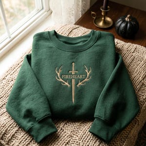 May include: A dark forest green crewneck sweatshirt featuring gold-coloured embroidery on the chest. The design depicts a central sword flanked by two deer antlers, with the word FIREHEART stitched in capital letters across the sword blade.