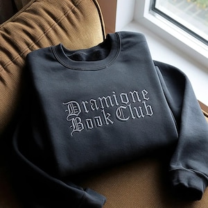 Op de afbeelding: Een antracietgrijze sweater met ronde hals en witte geborduurde tekst op de borst die Dramione Book Club spelt in een gotisch lettertype. Het kledingstuk heeft lange mouwen, een geribbelde halslijn en een zachte fleece voering.