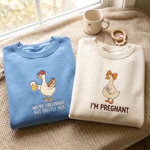 Könnte beinhalten: Ein blaues Sweatshirt mit einer gestickten Gans, die ein Bier und einen Hotdog hält, und dem Text WE'RE PREGNANT BUT MOSTLY HER, neben einem cremefarbenen Sweatshirt mit einer Gans mit Schleife und dem Text I'M PREGNANT.