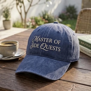 May include: A blue denim baseball cap featuring the embroidered white text "MASTER OF SIDE QUESTS" on the front panel. The cap has a structured crown, a curved brim, and stitched eyelets for ventilation, presenting a classic six-panel design.