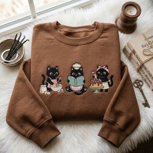 Könnte beinhalten: Ein braunes Sweatshirt mit Rundhalsausschnitt und einer Stickerei von drei backenden schwarzen Katzen. Eine Katze rührt Zutaten um, eine liest in einem Kochbuch und die dritte verziert einen Kuchen. Das Motiv zeigt Backutensilien, Eier und einen Kuchen.