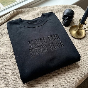 Può includere: Una felpa nera a girocollo con il testo ELDER EMO MUMS CLUB ricamato in filo nero sul petto con un carattere in stile gotico. Il capo presenta uno scollo a coste e maniche lunghe, presentato in un orientamento piegato e piatto.