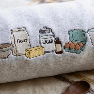 May include: A light gray fleece blanket featuring an embroidered design of baking ingredients including a mixing bowl with a spoon, a bag labeled flour, a stick of butter, a jar labeled sugar, a small bottle, a carton of eggs, and a measuring cup.