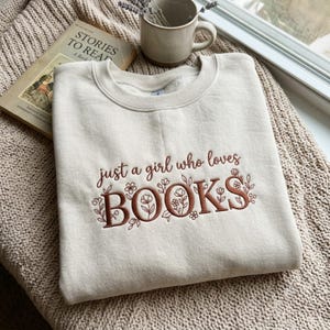 May include: A cream-colored crewneck sweatshirt featuring brown embroidered text that reads "just a girl who loves BOOKS" with delicate floral and vine illustrations woven between the letters, centered on the front of the garment.