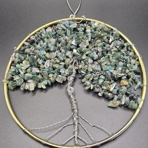 Può includere: Decorazione da parete artigianale dell'albero della vita. Il design presenta una cornice circolare color ottone con un albero costruito con filo d'argento. Le foglie dell'albero sono fatte di piccole pietre verdi e beige. Un anello di filo d'argento in alto per appendere.