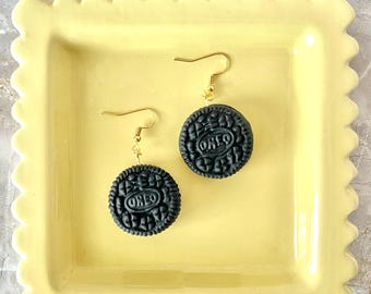 May include: A pair of dangle earrings featuring black circular charms embossed with the text OREO, suspended from gold-toned metal fishhook wires. The charms replicate the appearance of sandwich cookies with detailed textured edges.