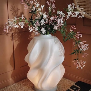 Op de afbeelding: Een witte, sculpturale vaas met een wervelend ontwerp, van binnenuit verlicht, bevat een boeket witte en roze bloemen. De vaas staat op een grijze voet, geplaatst op een getextureerd tapijt. De bloemen hebben groene bladeren.