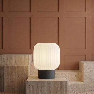 May include: A modern table lamp with a ribbed, off-white shade and a dark gray base. The lamp is illuminated, casting a soft glow. It sits on a stone-like surface, with a brown wall in the background. A white cord extends from the lamp.
