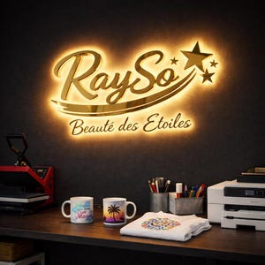 May include: A gold-colored sign with the text "RaySo Beauté des Etoiles" is illuminated on a dark wall. Two mugs, a heat press, and a printer sit on a wooden desk. A white t-shirt with a colorful design is also on the desk.