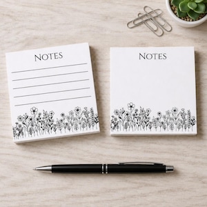Puede incluir: Dos blocs de notas cuadrados blancos con la palabra NOTES impresa en la parte superior. Un bloc tiene líneas horizontales y el otro está en blanco. Ambos tienen una ilustración de flores silvestres en línea negra en la base. Incluye un bolígrafo negro.