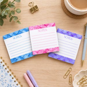 May include: Three square sticky note pads with white centres and lined paper. The top and bottom edges feature a watercolour quatrefoil pattern in blue, pink, and purple. Each pad has the word NOTES printed in the centre top area in coloured ink.