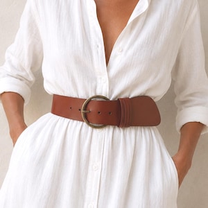 Puede incluir: Un vestido camisero blanco con botones, mangas largas y cuello. El vestido cuenta con un cintur&oacute;n ancho de cuero marr&oacute;n con una hebilla redonda de tono lat&oacute;n ajustado a la cintura. La tela tiene una apariencia texturizada y el vestido incluye bolsillos laterales.