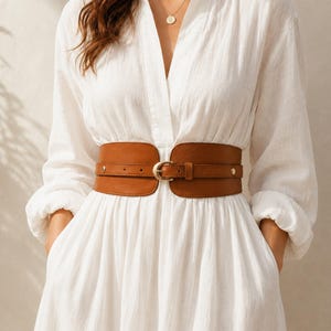 May include: A long-sleeved white dress with a V-neckline and a wide, tan-colored faux leather corset-style belt. The belt features a central gold-toned buckle, a thin strap overlay, and small gold-toned metal studs on each side of the front closure.